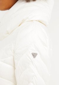 Vit puffjacka med quiltat mönster, huva och ett dekorativt triangulärt detaljer med rhinestones på vänster arm.