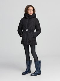 Czarna kurtka puchowa z wysokim kołnierzem, wcięciem w talii i srebrną klamrą paska. Noszona z czarnymi legginsami i granatowymi gumowcami.