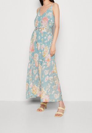 Kvinde iført blå, blomsterprintet, ærmeløs maxikjole med bælte og gule høje hæle sandaler, stående mod en ensfarvet hvid baggrund.
