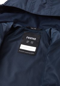 Interno della giacca blu navy con rivestimento in rete. L'etichetta nera mostra il nome del marchio "reima", la taglia "104" e i simboli per la manutenzione.