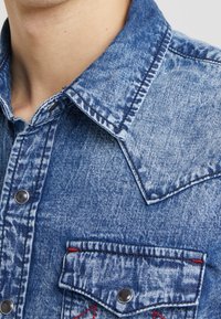 Ljusblå denimskjorta med spetsig krage, tryckknappar och två bröstfickor med röda detaljer och kontrasterande sömmar.