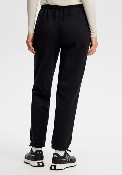 Osoba ubrana w czarne spodnie dresowe z elastycznym pasem, białe skarpetki, czarno-białe trampki oraz beżową bluzkę z długim rękawem, zwrócona tyłem.