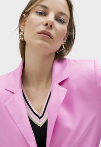 Pink blazer with structured lapels, worn over a black V-neck sweater featuring multicolored stripes. Gold hoop earrings with text detail.
