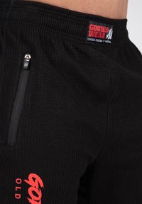 Black textured pants with a ribbed pattern, featuring a zippered pocket and a red logo label reading "GORILLA WEAR."