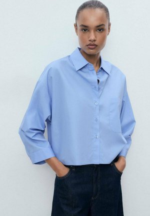 Light blue button-up shirt with a collar, single chest pocket, and three-quarter length sleeves, paired with dark denim jeans.