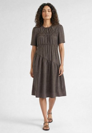 Femme aux cheveux bouclés portant une robe marron longueur genou avec des manches courtes et des sandales, debout devant un fond clair uni.