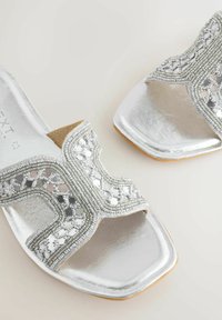 Mules ouvertes argentées à bout carré, ornées de motifs décoratifs complexes en perles et miroirs sur les brides.