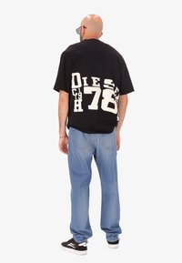 Uomo con testa rasata che indossa una maglietta nera con grafica, jeans azzurri chiari, sneakers nere e occhiali da sole, in piedi di spalle su sfondo bianco.