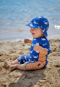Traje de baño azul con patrones de cangrejos, de mangas cortas y pantalones cortos a rayas naranjas. Sombrero azul a juego con protección para el cuello. Entorno arenoso.