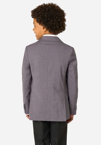 Dziecko z kręconymi włosami, ubranym w szary żakiet i czarne spodnie, stoi plecami do obiektywu na jednolitym tle.