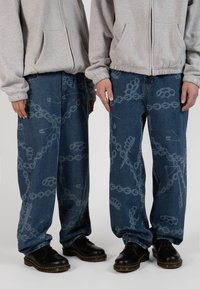 Vaqueros de mezclilla de corte holgado, con un sutil patrón gráfico en azul claro de cadenas y herramientas, combinados con zapatos negros de cordones.