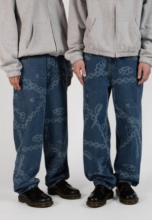 Jean en denim à coupe décontractée, avec un motif graphique léger de chaînes et d'outils en bleu clair, associée à des chaussures noires à lacets.