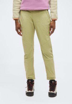 Persoon die beige outdoorbroeken en beige wandelschoenen met zwarte zolen en rode veters draagt, staand tegen een effen achtergrond.