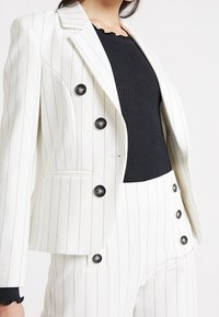Bílý blazér s proužky a černými knoflíky, nošený přes černý žebrovaný top. Střižený design se striktními rameny a štíhlým střihem.