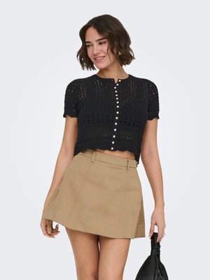 Joven mujer con un top corto de ganchillo negro con botones y una falda beige en línea A, sujetando un bolso negro a su lado.