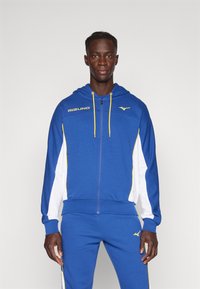 Hanorac bleumarin cu fermoar, accente galbene și panouri laterale albe, fabricat dintr-un material moale. Include șnururi și logo pe piept. Pantaloni asortați bleumarin.