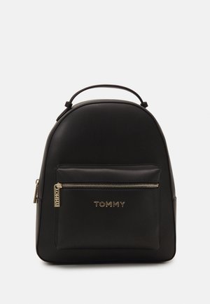 Sort læderrygsæk med øverste håndtag, frontlomme med lynlås, guldlynlås og "TOMMY" logo på lommen mod ensfarvet baggrund.