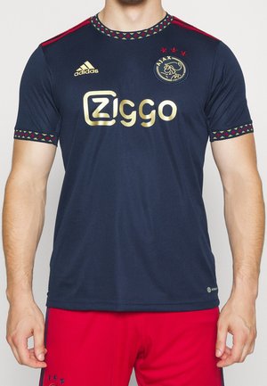 Man draagt een donkerblauw Ajax-voetbalshirt met gouden sponsorlogo en rode korte broek, staand tegen een effen achtergrond.