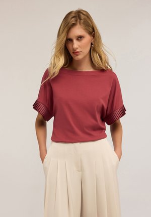 Camicia bordeaux a maniche corte con accentuazioni plissettate. Abbinata a pantaloni crema a vita alta, che mostrano un taglio rilassato e una struttura ben definita.