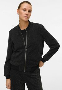 Veste bomber noire avec fermeture éclair à l'avant et poignets côtelés. Fabriquée en tissu texturé avec un subtil éclat. Un pantalon assorti est visible.