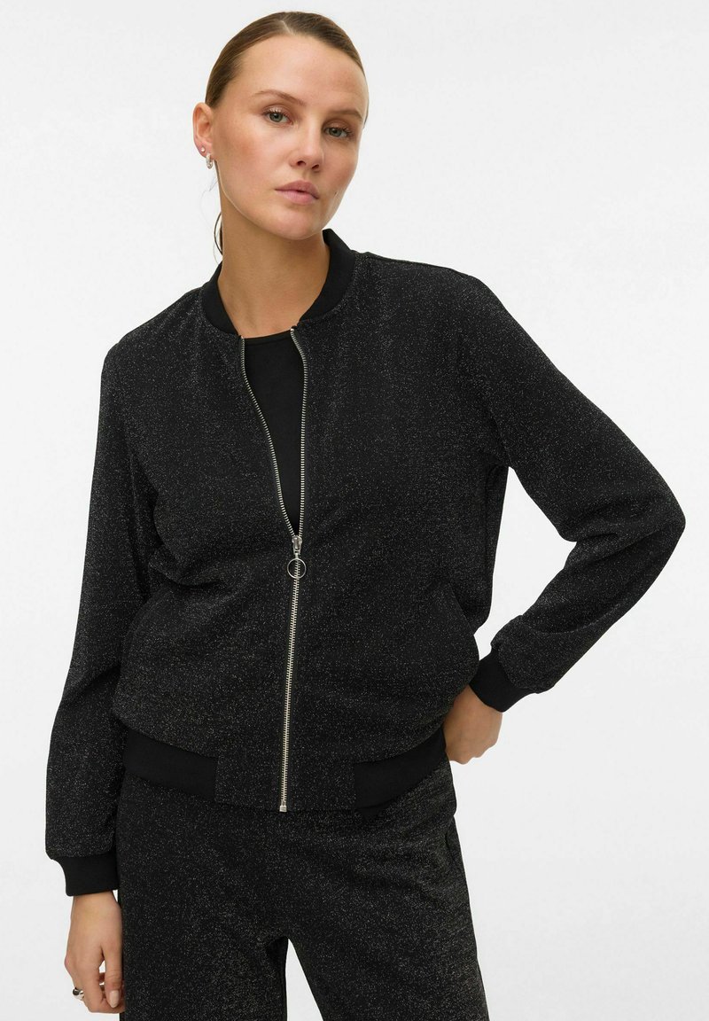 Veste bomber noire avec fermeture éclair à l'avant et poignets côtelés. Fabriquée en tissu texturé avec un subtil éclat. Un pantalon assorti est visible.