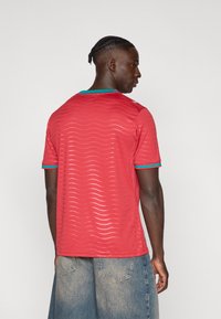 Κόκκινο κοντομάνικο μπλουζάκι με κυματιστό μοτίβο υφής, με μπορντούρα στο λαιμό σε μπλε-πράσινο χρώμα, φορεμένο με μπλε τζιν σορτς, από πίσω.