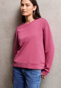 Sweatshirt rose à manches longues avec un col rond, ornée de petites clous sur les coutures des épaules, associée à un jean bleu sur un fond en béton.
