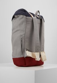 Hallitus hallist kangast seljakot, millel on tume sinine ülaosa ja punane alaosa, reguleeritavad beežid rihmad, külgmine luku tasku ja tugevdusõmblused rihmadel.