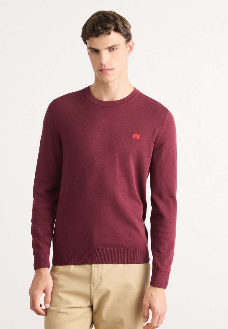 Jeune homme portant un pull marron ajusté avec un petit logo rouge sur la poitrine et un pantalon beige, se tenant contre un fond blanc.