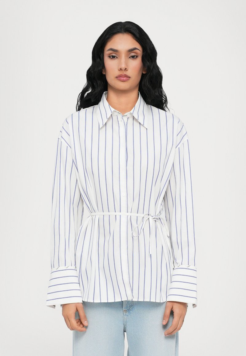 Camicia bianca con righe verticali blu, maniche lunghe e vita con cintura. Presenta polsini larghi e colletto classico.