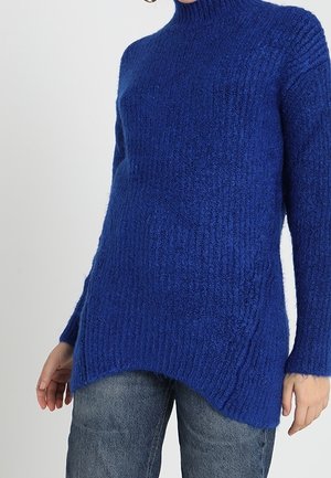 Femme portant un pull long, texturé bleu roi avec un col montant, associé à un jean bleu décontracté.