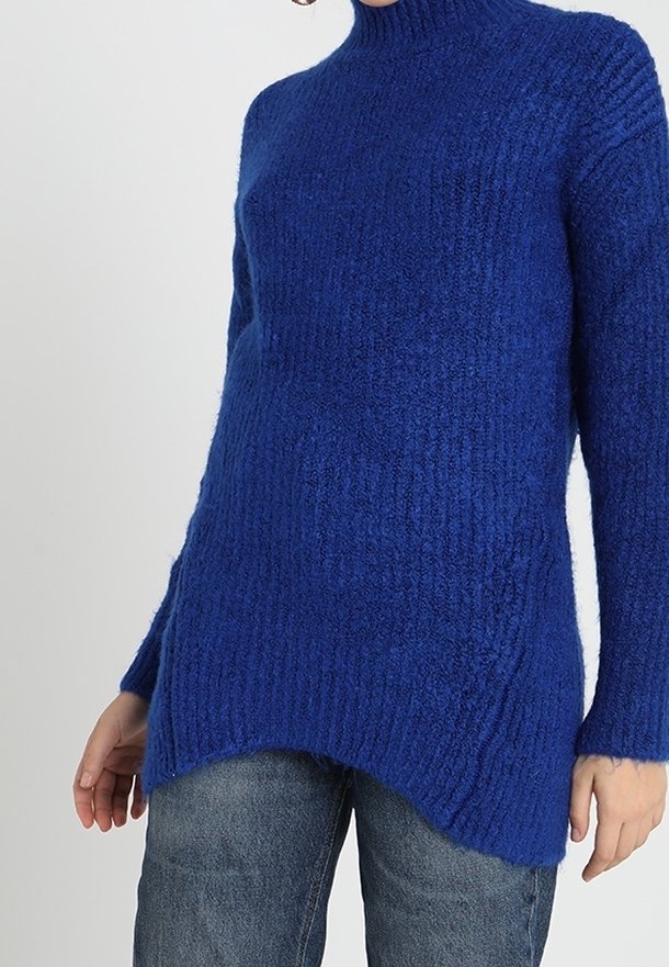 Femme portant un pull long, texturé bleu roi avec un col montant, associé à un jean bleu décontracté.