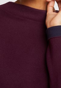 Burgunderfarbener Pullover mit geripptem Muster und rundem Ausschnitt; verfügt über kontrastierende dunkelblaue Bündchen als Detail. Weicher, sichtbarer Strickstoff.