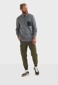 Pullover grigio lavorato a maglia con tasca nera, abbinato a pantaloni cargo oliva con tasche laterali e sneaker nere con accenti crema.
