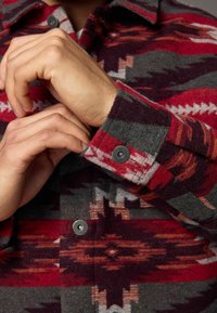 Chemise en flanelle à motif rouge et gris avec des designs géométriques, des poignets à boutons et une texture douce. Main visible ajustant la manche.