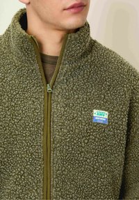 Chaqueta de forro polar en verde olivo con un cuello alto, cierre de cremallera y superficie texturizada. Presenta un parche de marca azul y verde en el pecho.