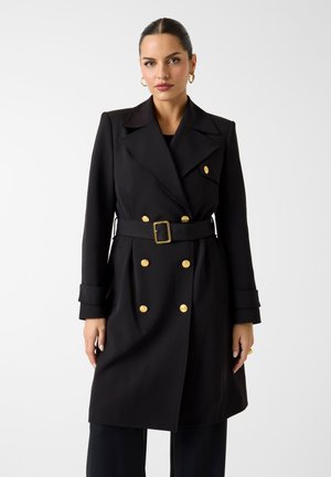 Femme portant un manteau croisé noir avec des boutons dorés et une ceinture noire assortie, debout devant un fond blanc uni.