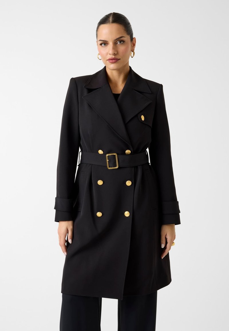 Femme portant un manteau croisé noir avec des boutons dorés et une ceinture noire assortie, debout devant un fond blanc uni.