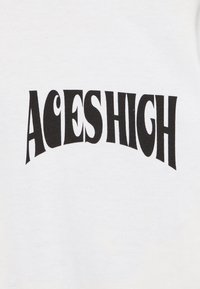 Biały materiał z wyraźnym czarnym tekstem "ACES HIGH" o zakrzywionym kształcie. Czcionka jest zabawna i lekko zniekształcona.