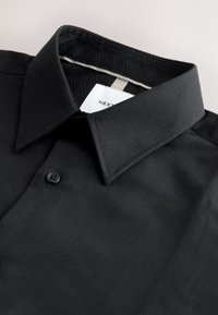 Chemise noire à col avec tissu texturé et un seul bouton visible, montrée pliée avec une étiquette blanche à l'intérieur du col.