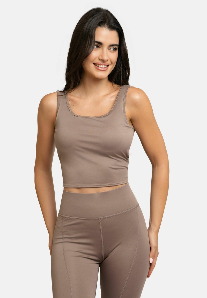Femme aux longs cheveux foncés portant un crop top sans manches taupe ajusté assorti et des leggings taille haute, souriante et regardant sur le côté.