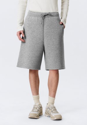 Persoon draagt lichtgrijze knielange shorts met trekkoord, wit shirt met lange mouwen, beige sokken en beige sneakers, staand met handen in de zakken.