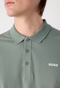 Salve roheline polo särk, millel on krae, kolm nööpaukku ja valge brodée 'HUGO' logo rinnal. Tekstuuriline kangas.
