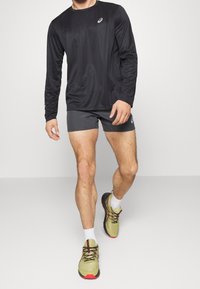 Schwarzes Langarm-Sportshirt, kombiniert mit grauen Shorts und grünen Turnschuhen mit schwarzen Akzenten und roten Sohlen, getragen mit weißen Socken.