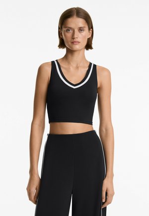 Model nosi brezrokavnik črn crop top z belim robom in visoko pasom črne hlače, pozira ob enobarvni beli steni.