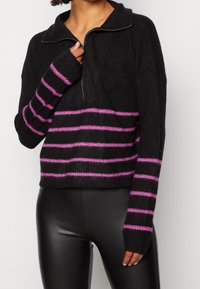 Schwarzer Strickpullover mit Reißverschlusskragen, der rosa und schwarze horizontale Streifen aufweist. Enganliegende Ärmel mit Daumenlöchern. Strukturierte Oberfläche.