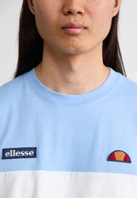 Světle modré tričko s krátkým rukávem a bílou spodní částí. Obsahuje vyšívané logo "ellesse" a barevně různorodý akcent loga na prsou.