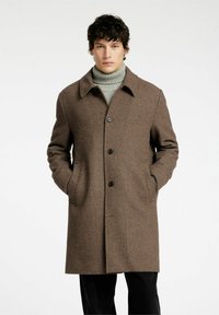 Cappotto a motivo houndstooth marrone con colletto a punte, chiusura frontale a bottoni e tasche laterali. Indossato sopra un maglione a collo alto grigio.