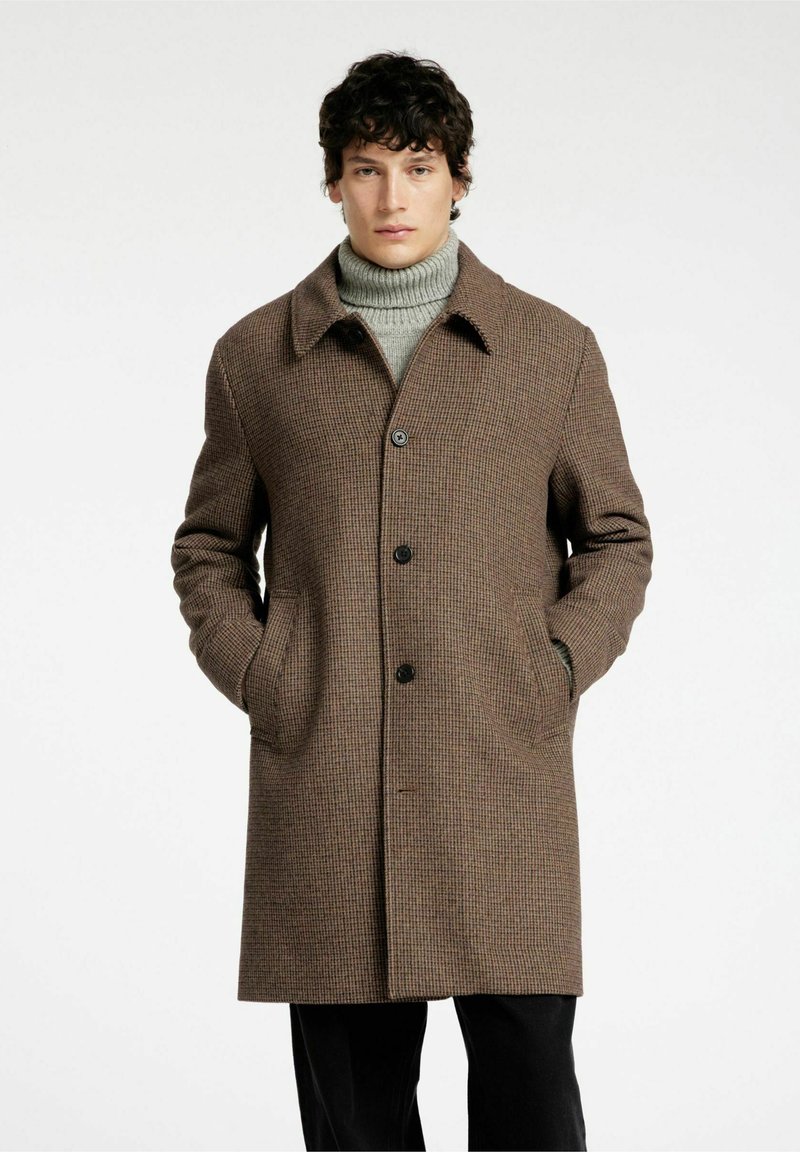 Cappotto a motivo houndstooth marrone con colletto a punte, chiusura frontale a bottoni e tasche laterali. Indossato sopra un maglione a collo alto grigio.