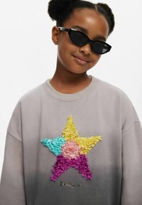 Grauer Pullover mit einem mehrfarbigen, strukturierten Stern-Design aus übereinanderliegenden Stofflagen. Akzentuiert mit einem kleinen, funkelnden Logo unter dem Stern.